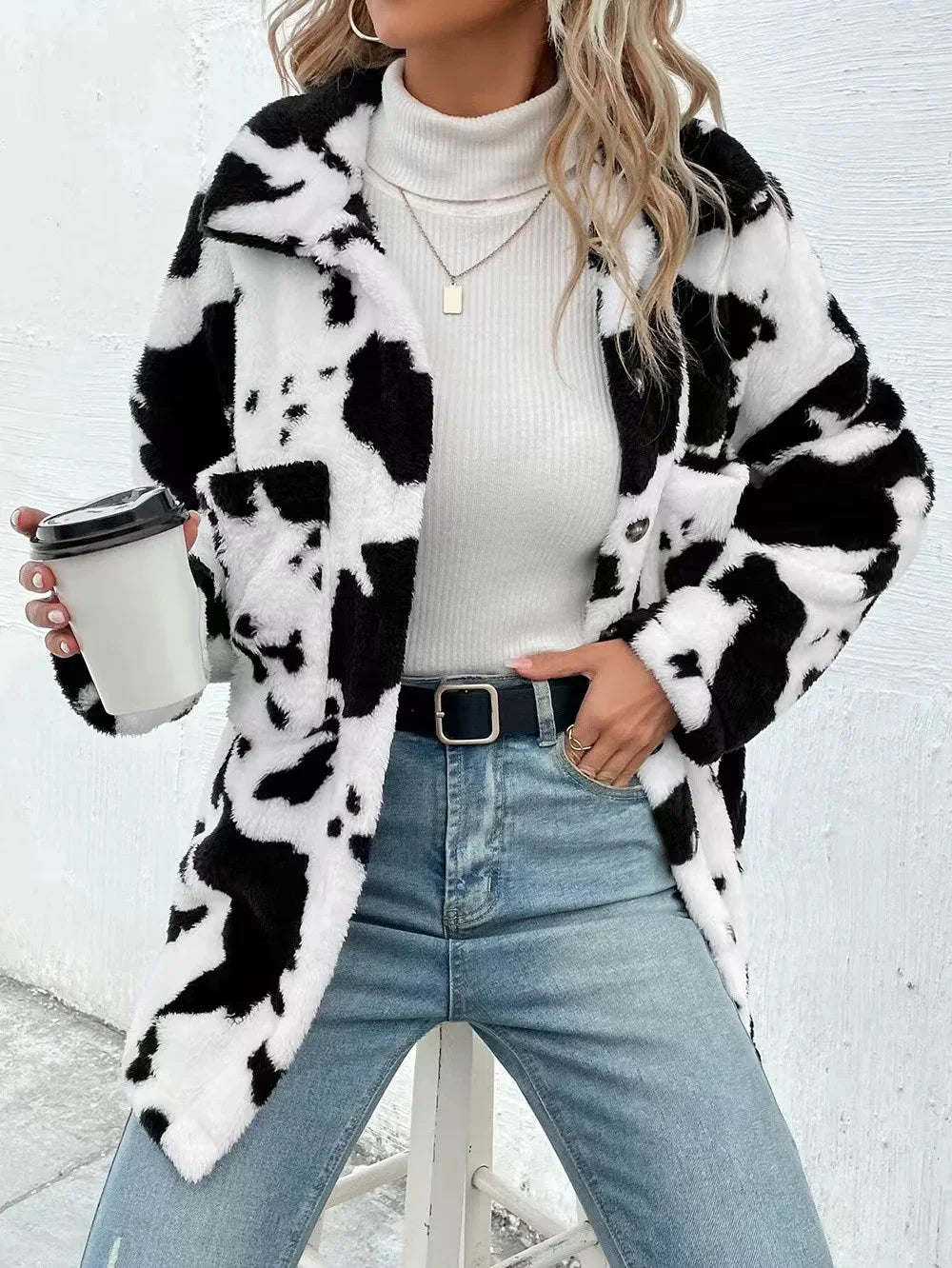 Woman wearing black and white cow print fur jacket, white turtleneck, blue jeans, holding coffee cup