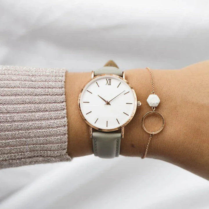 Minimalist wristwatch with white face, gold case, gray leather strap, worn with delicate gold bracelet