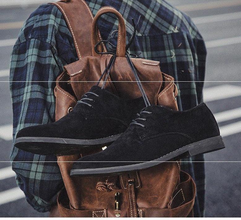 Black suede casual shoes hanging on brown leather backpack worn by person in green plaid shirt