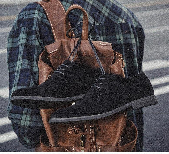 Black suede casual shoes hanging on brown leather backpack worn by person in green plaid shirt