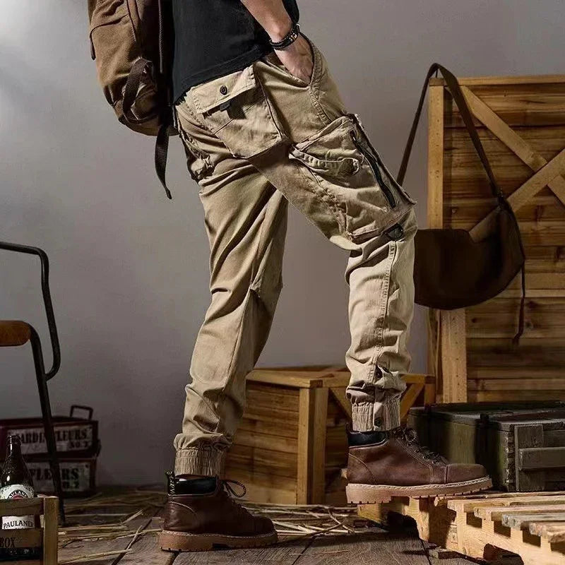 Man wearing beige cargo jogger pants and brown boots standing indoors with wooden crates and backpack