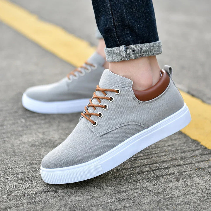Close-up of light gray casual sneakers with brown laces and white soles worn with rolled-up dark jeans on pavement