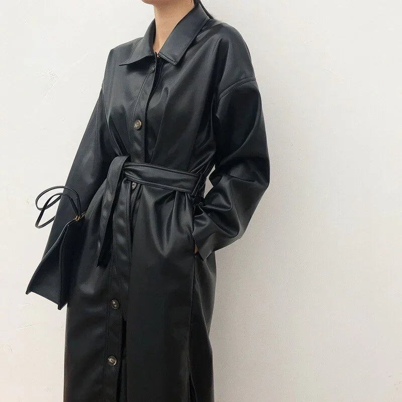 Woman wearing a black belted faux leather trench coat with buttons, holding a matching black handbag