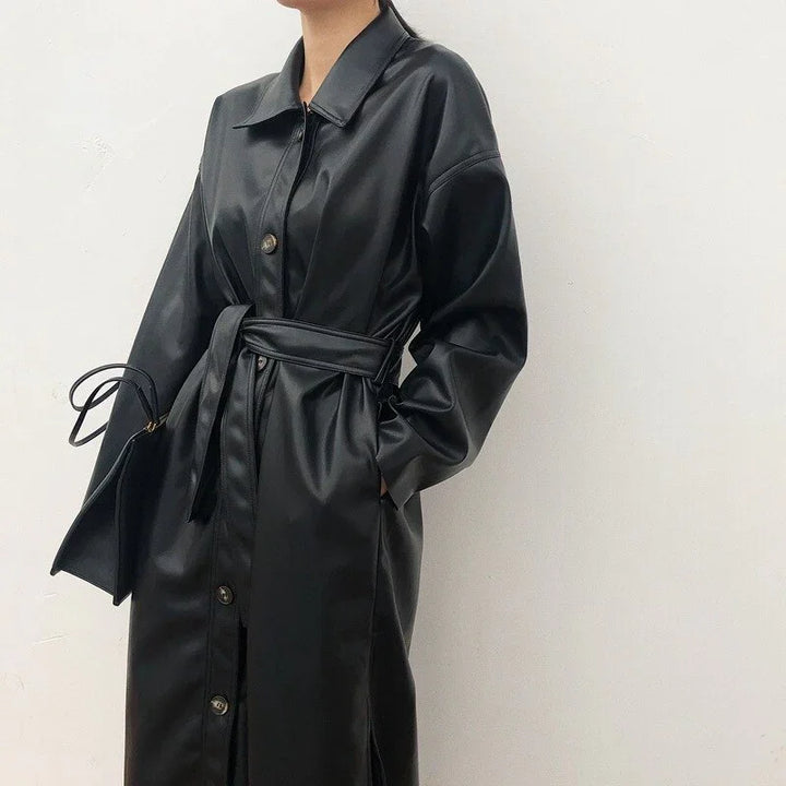 Woman wearing a black belted faux leather trench coat with buttons, holding a matching black handbag