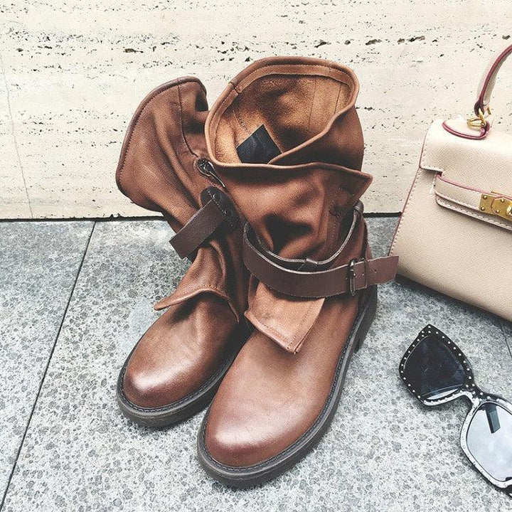 brown leather boots with buckle detail on stone floor next to beige handbag and black studded sunglasses