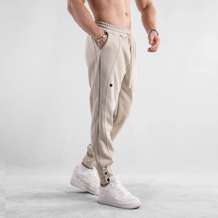 Male model wearing beige jogger pants with side seams and white designer sneakers on gray background
