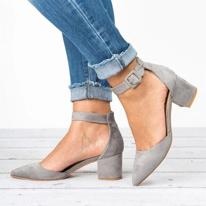 Grey suede block heel sandals with ankle strap worn with rolled-up blue jeans on white floor