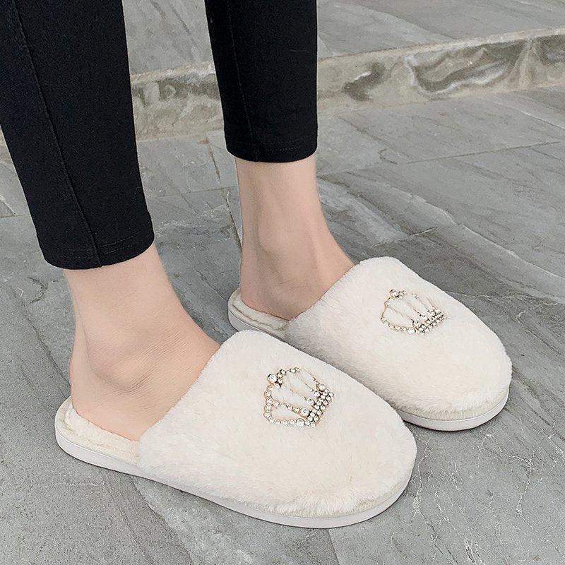 Fuzzy white slippers with decorative pearl crown emblem worn with black pants on gray stone floor