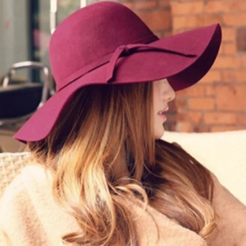 Woman wearing a large purple floppy hat with bow detail indoors by brick wall