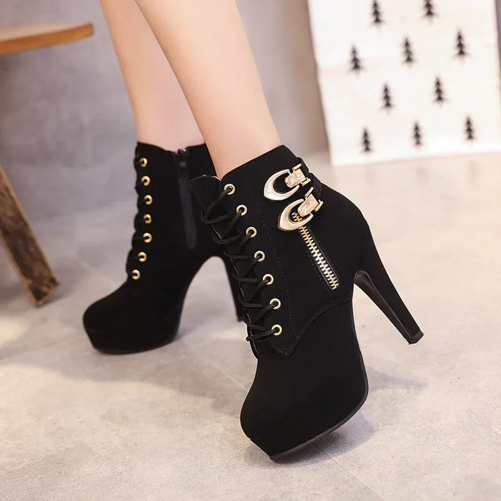 Black high heel ankle boots with lace-up front, gold zipper, and buckle details on gray floor
