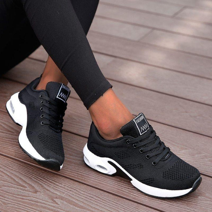 Black fashion sneakers with white sole and mesh design worn with black leggings on wooden floor