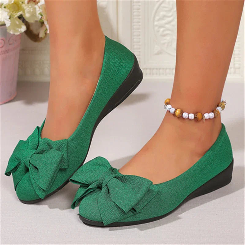 Green flat shoes with large bows worn with beaded ankle bracelet on gray floor