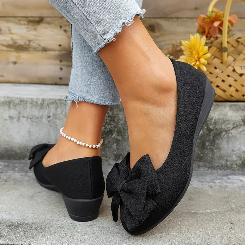 Black wedge shoes with large front bow, worn with frayed hem jeans and pearl ankle bracelet on concrete