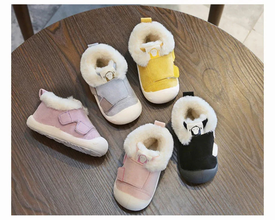 Five pairs of soft baby shoes with faux fur lining in pink, gray, yellow, black, and pale pink on a wooden table