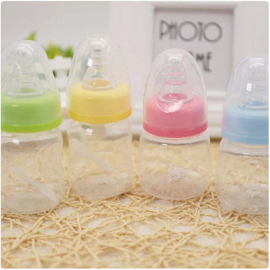 Four small baby bottles with clear nipples and colored caps in green, yellow, pink, and blue on woven mat