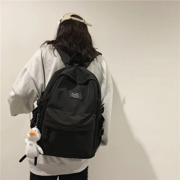 Person wearing gray sweatshirt and beige cap with black backpack and white duck keychain against plain wall
