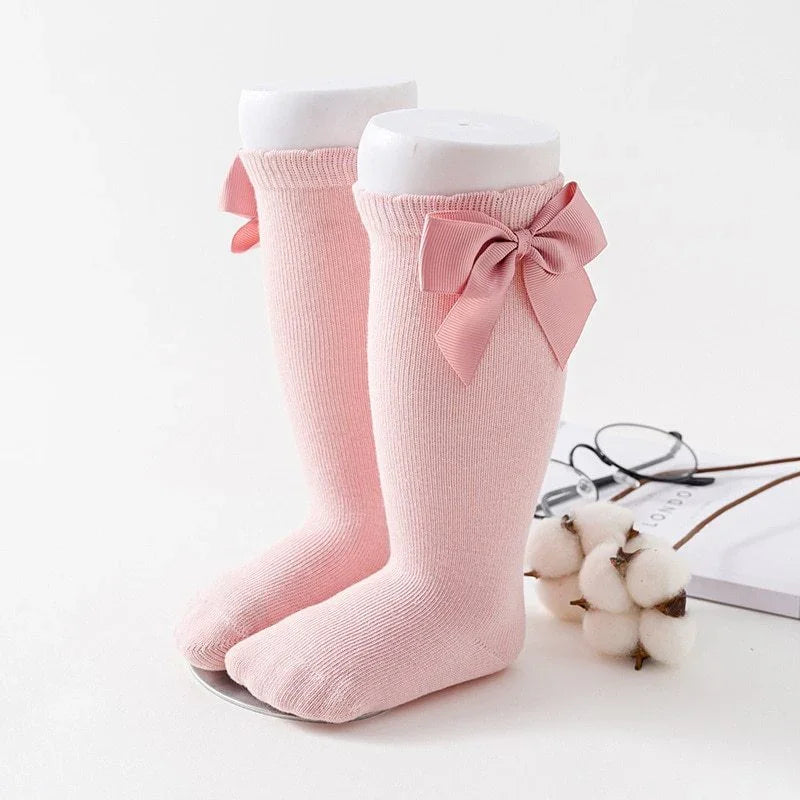 Pair of pink baby socks with large bows on a white display stand with cotton flowers and glasses nearby