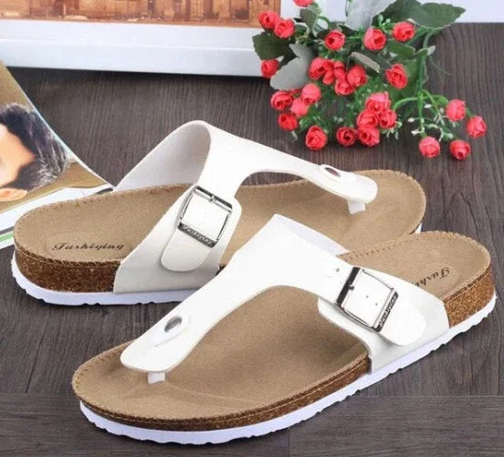 White leather thong sandals with adjustable silver buckles and cork footbeds on wooden floor