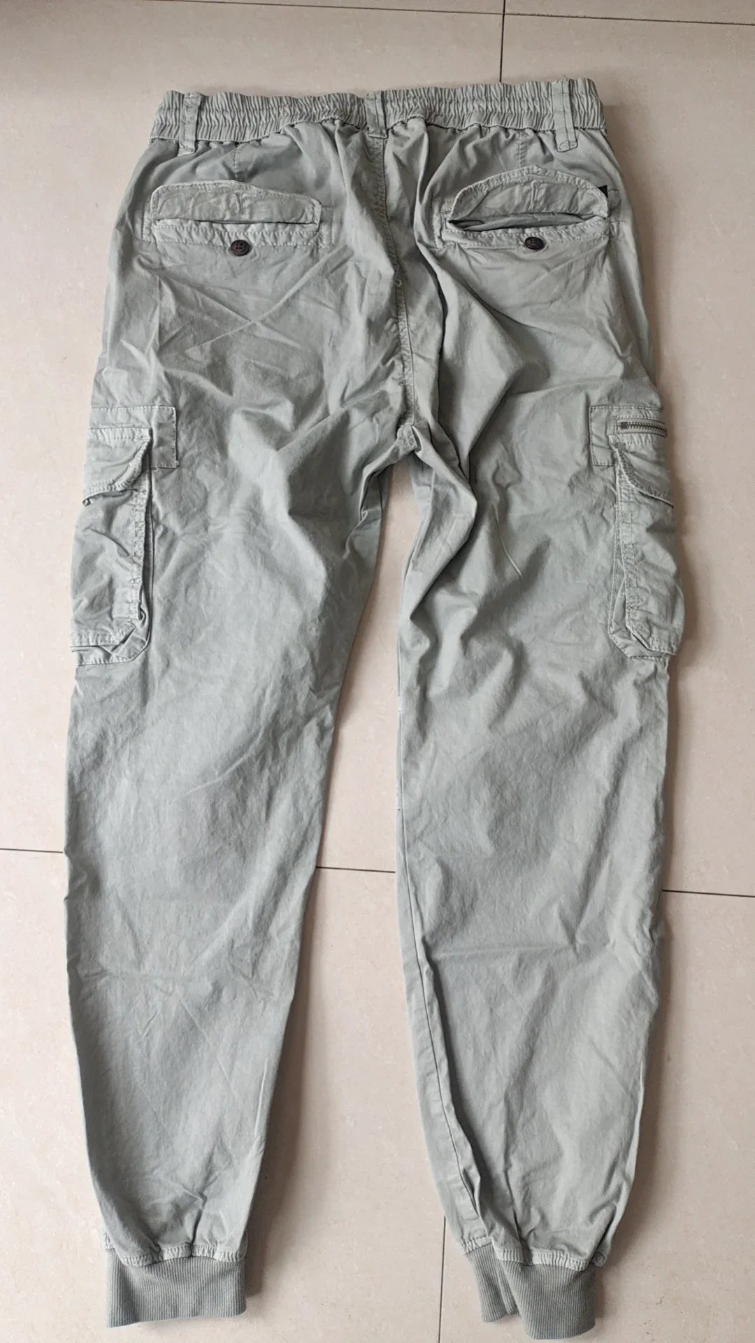 Light grey cargo jogger pants with elastic waistband, buttoned back pockets, and cuffed ankles