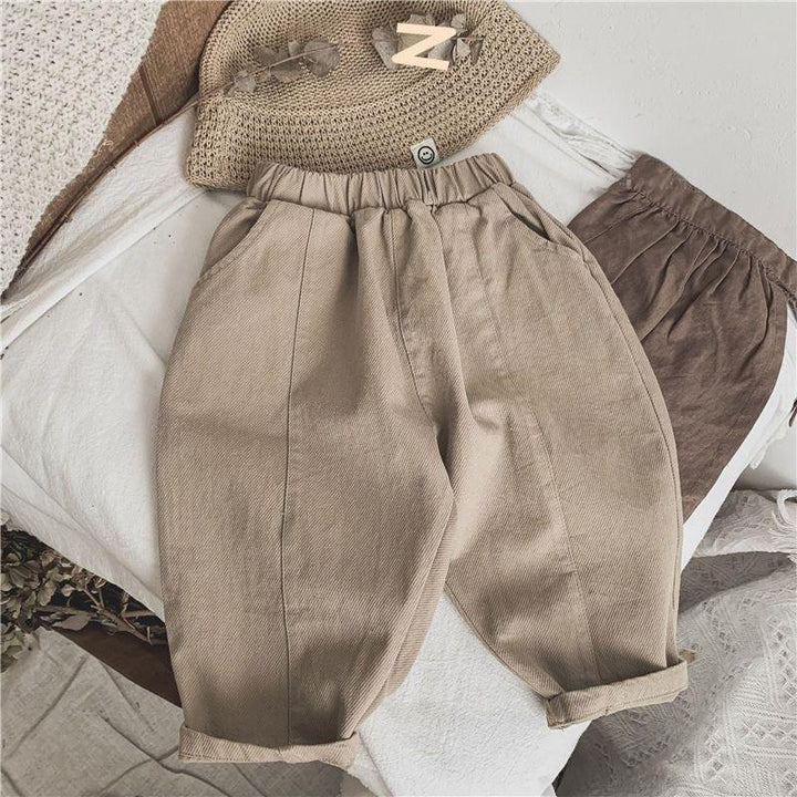 Beige cotton toddler pants with elastic waistband and pockets on white textured fabric with brown pants and a woven hat