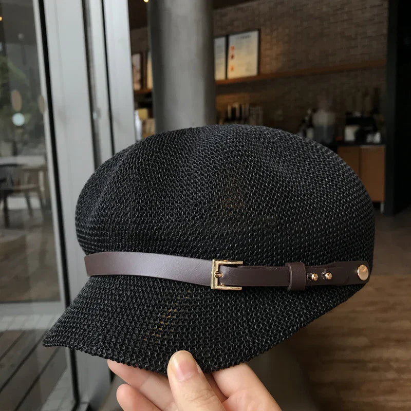 Hand holding black woven summer hat with brown leather strap and gold buckle in cafe setting