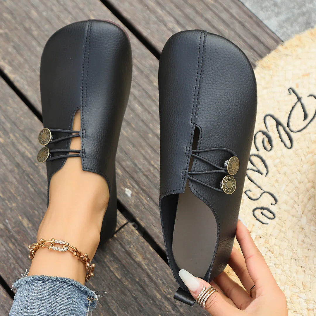 Black leather slip-on flats with round metal button accents, worn on wooden floor with denim jeans and anklet