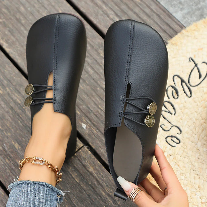 Black leather slip-on flats with round metal button accents, worn on wooden floor with denim jeans and anklet