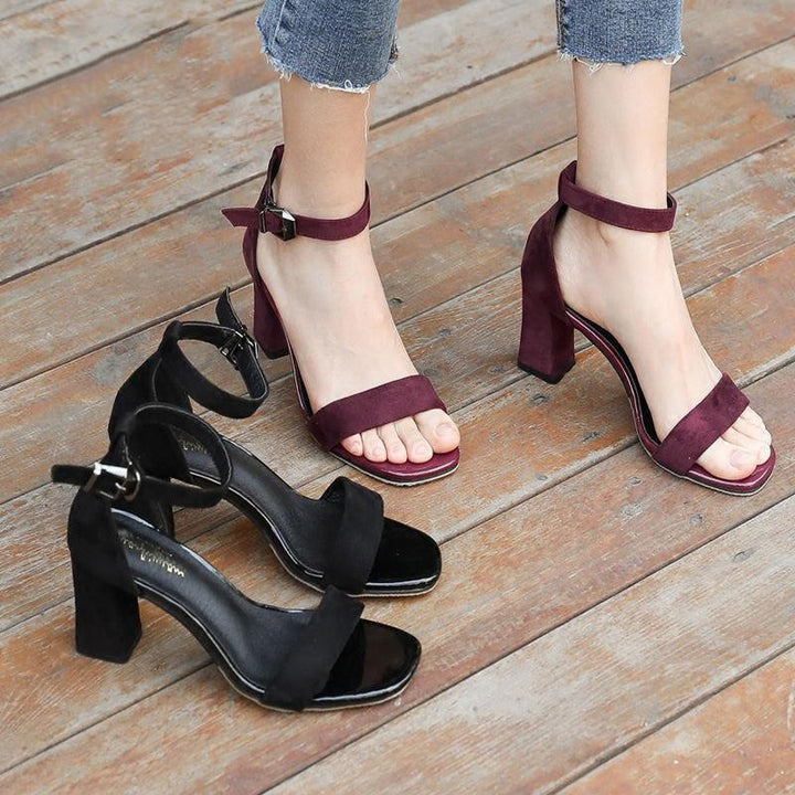 Woman wearing burgundy suede ankle strap block heel sandals with black suede pair on wooden floor