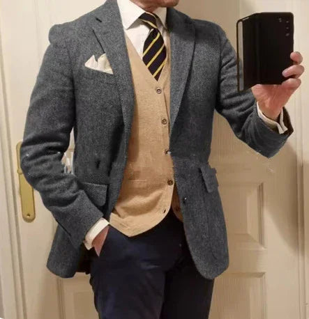 Man wearing grey tweed blazer, beige cardigan, white shirt, striped tie, and navy pants taking mirror selfie