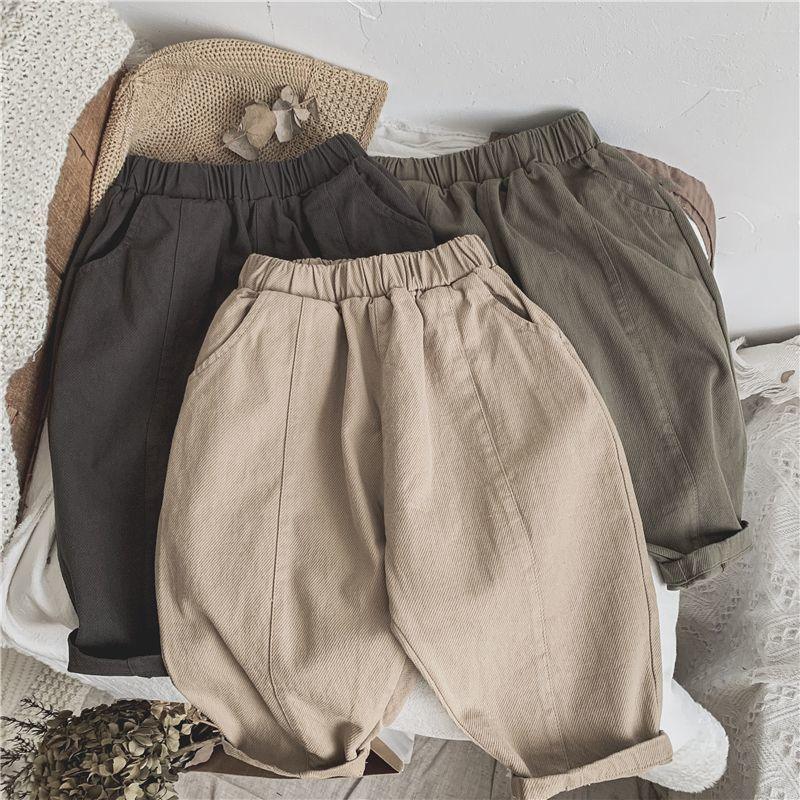 Three pairs of casual elastic waist pants in beige, olive green, and dark gray laid on a textured surface with dried leaves and knit fabric