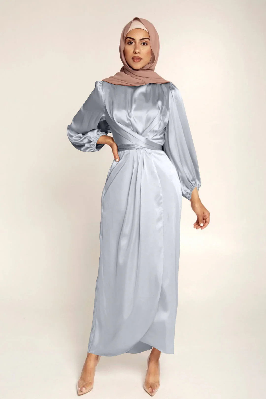 Woman wearing light blue satin long dress with balloon sleeves and beige hijab, posing against plain background