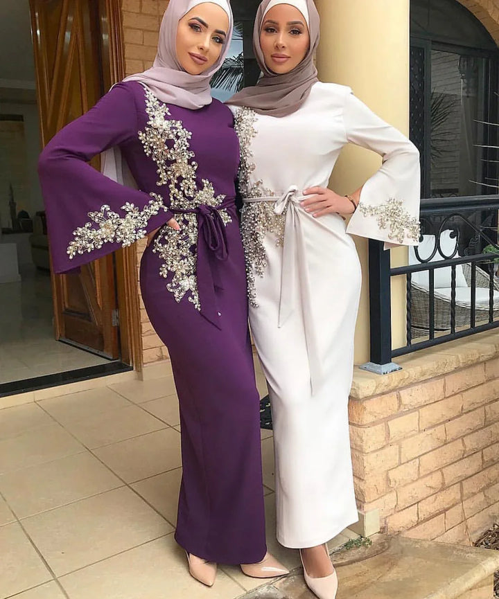Two women wearing elegant modest abaya dresses with pearl and floral embroidery, one in purple and one in white