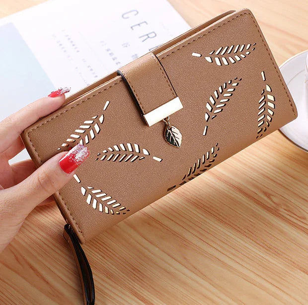Brown women's wallet with leaf cutout design and leaf charm, held by hand with red glittery nails on wooden surface