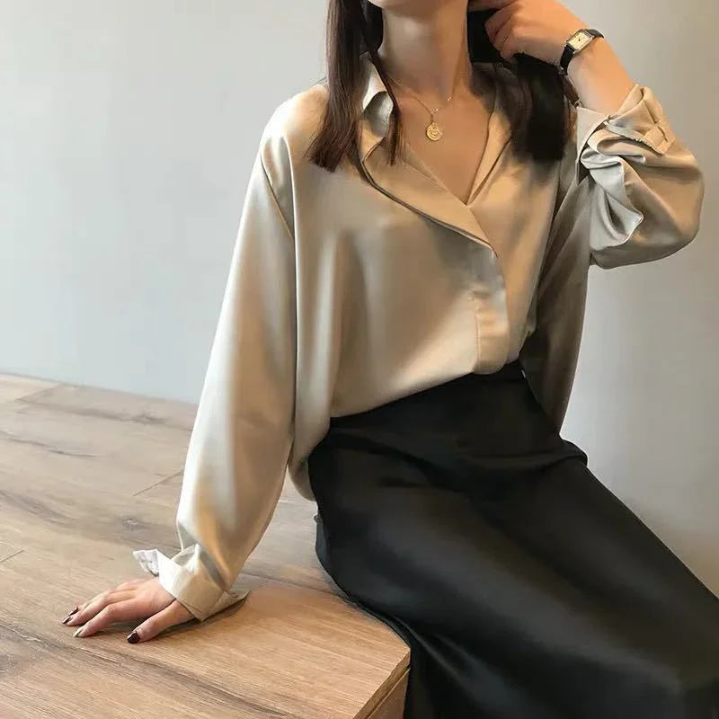 Woman wearing beige satin blouse and black silk skirt sitting on wooden table in minimalist room