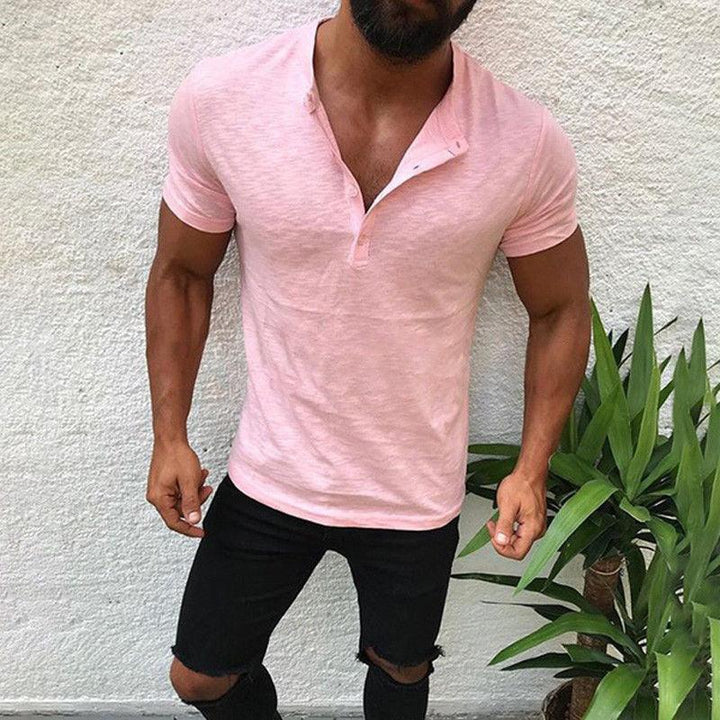 Muscular man wearing a light pink short sleeve henley shirt and ripped black jeans against a white wall with green plants