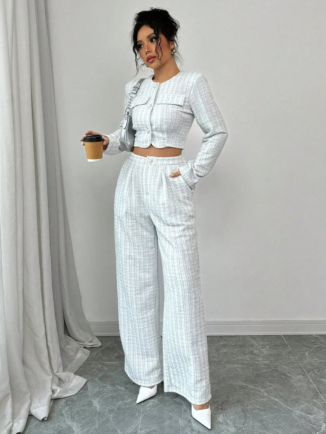 Woman in white textured crop jacket and wide-leg pants holding coffee cup in minimalist room