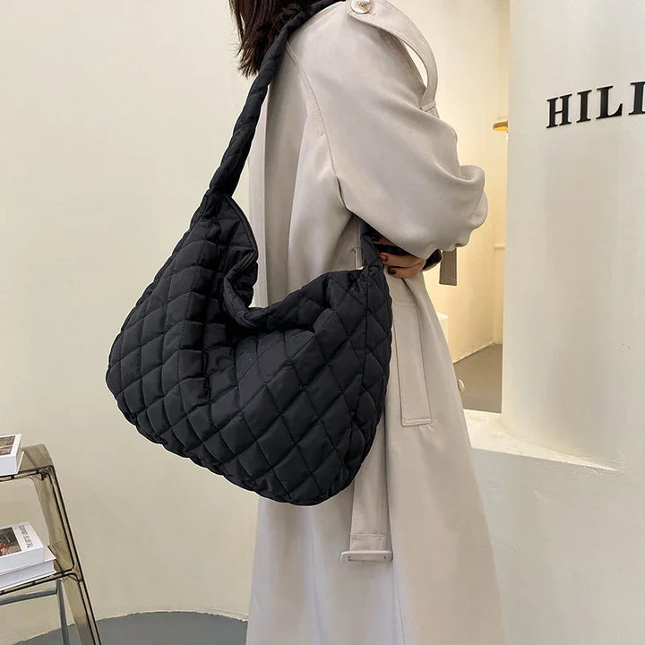 Woman wearing beige trench coat carrying large black quilted shoulder bag in minimal interior