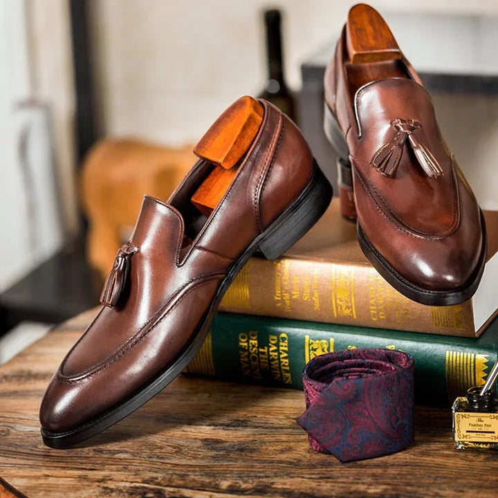 Brown leather tassel loafers displayed on vintage books with a paisley pocket square on wooden table