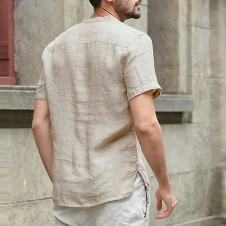 Man wearing beige short-sleeve linen shirt and light pants standing against stone wall
