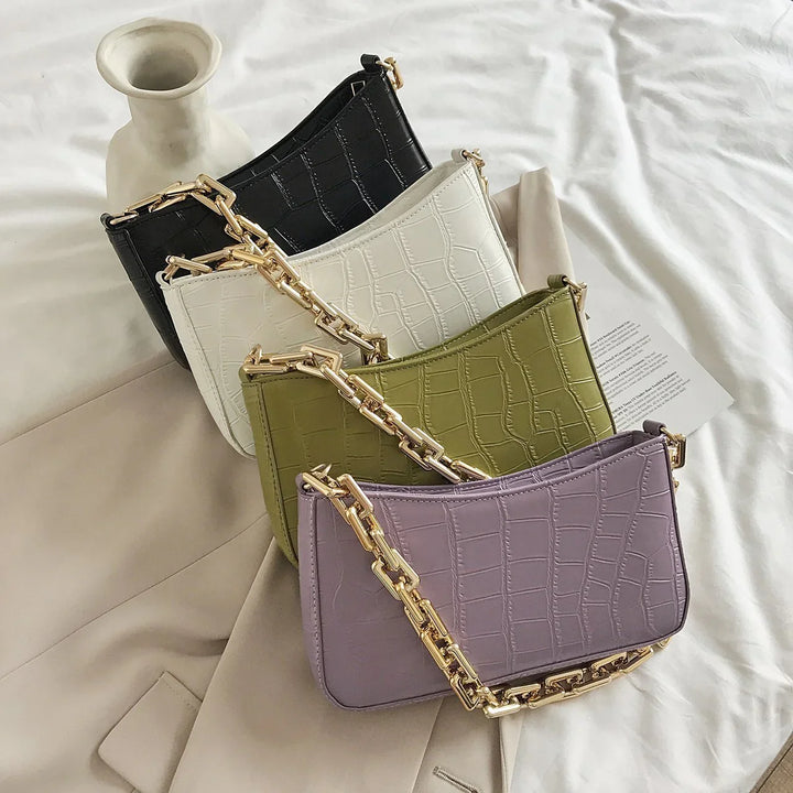 Four small crocodile-embossed leather handbags in black, white, green, and purple with gold chain straps on beige fabric