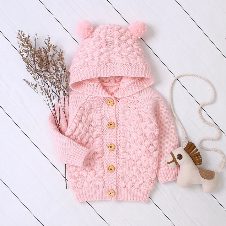 Pink knitted baby cardigan with pompom hood, wooden buttons, dried flowers, and toy horse on white floor