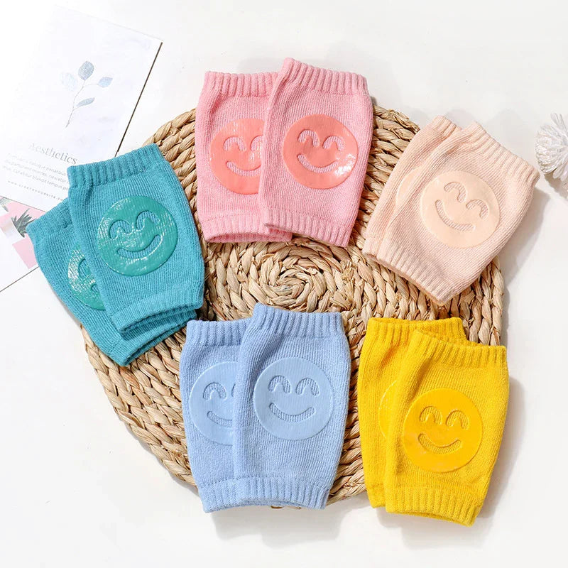 Colorful knit baby knee pads with smiley face grips in pink, blue, yellow, green, and beige on woven mat