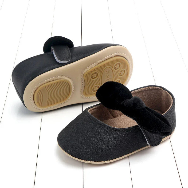 Black baby soft sole shoes with non-slip beige rubber soles and bow detail on white wooden floor