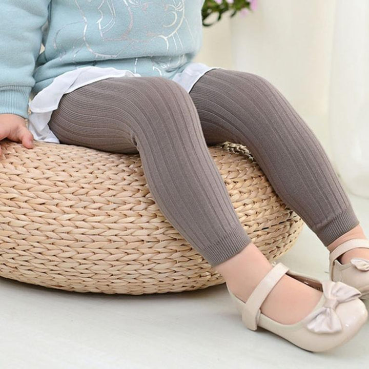 Toddler wearing brown ribbed leggings and beige bow shoes sitting on woven ottoman