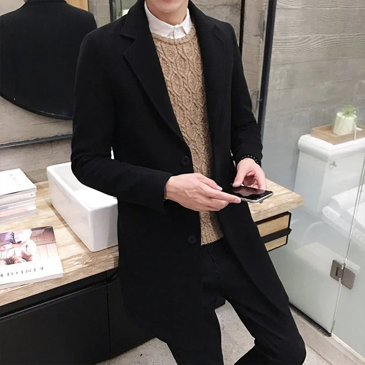 Man wearing black overcoat and brown sweater holding smartphone in modern bathroom with marble countertop