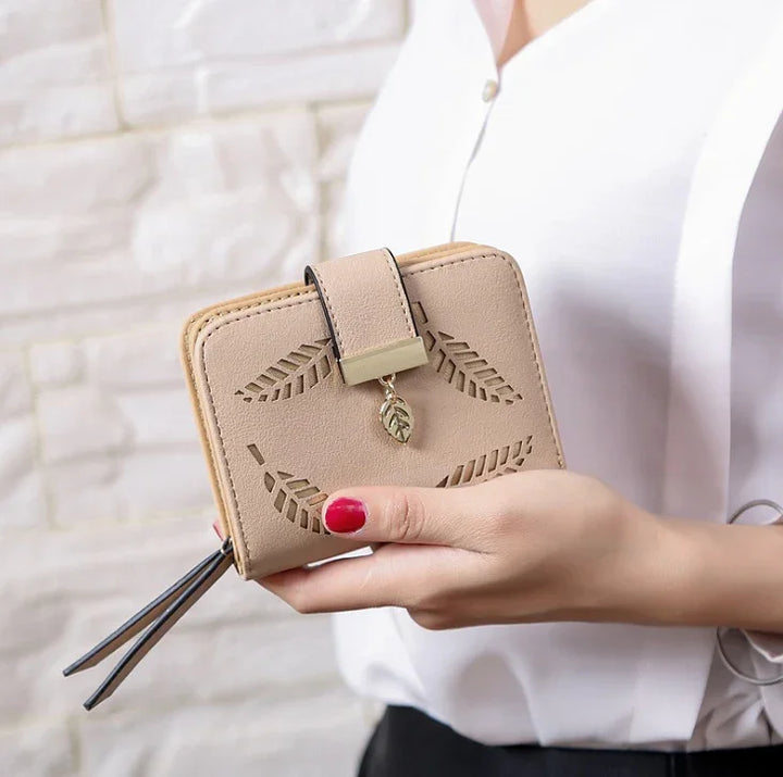 Beige leather wallet with leaf cutout design and gold leaf charm held by person with red nails