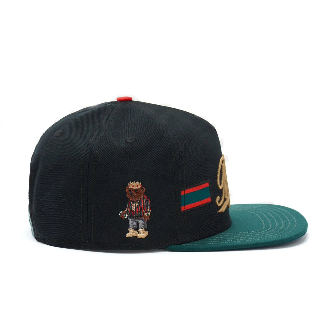 Black snapback hat with green brim, embroidered bear in striped shirt and gold crown detail