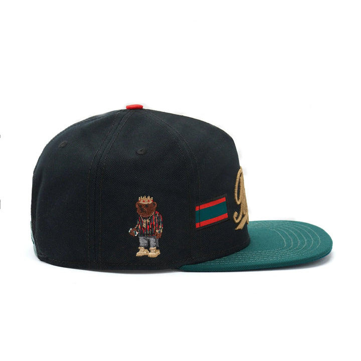 Black snapback hat with green brim, embroidered bear in striped shirt and gold crown detail