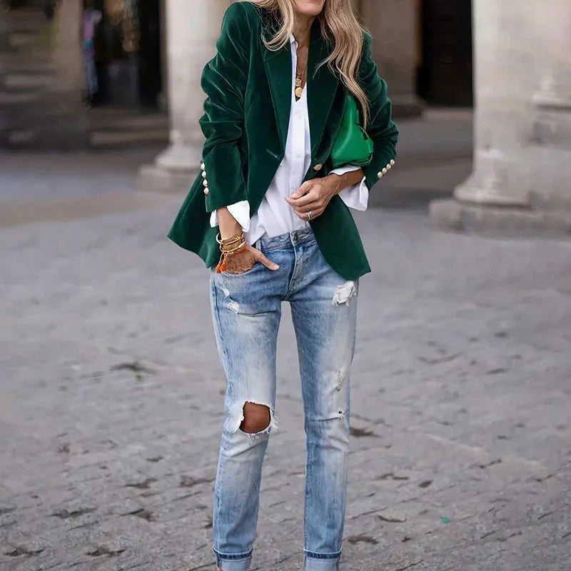 Woman wearing green velvet blazer, white blouse, ripped light blue jeans, and green handbag on city street