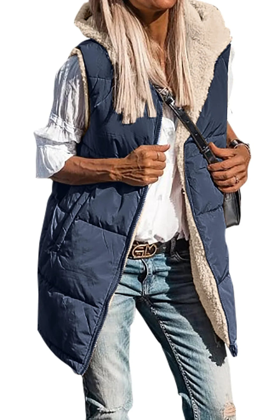 Woman wearing navy blue sleeveless puffer vest with beige fleece lining, white blouse, and ripped jeans